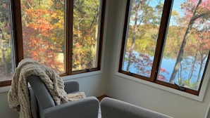 A corner room with two grey armchairs, a large window with a view of autumn trees and a lake.