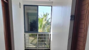 Un estrecho pasillo con suelo de baldosas blancas, una puerta corredera de vidrio que da a un balcón con vistas a la vegetación, y puertas de madera a ambos lados.