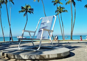 A beach chair with a striped backrest and metal frame, positioned on a concrete platform with palm trees and the ocean in the background. - Apê na Ponta Verde com Wi-Fi e Piscina no Topo (Maceió)