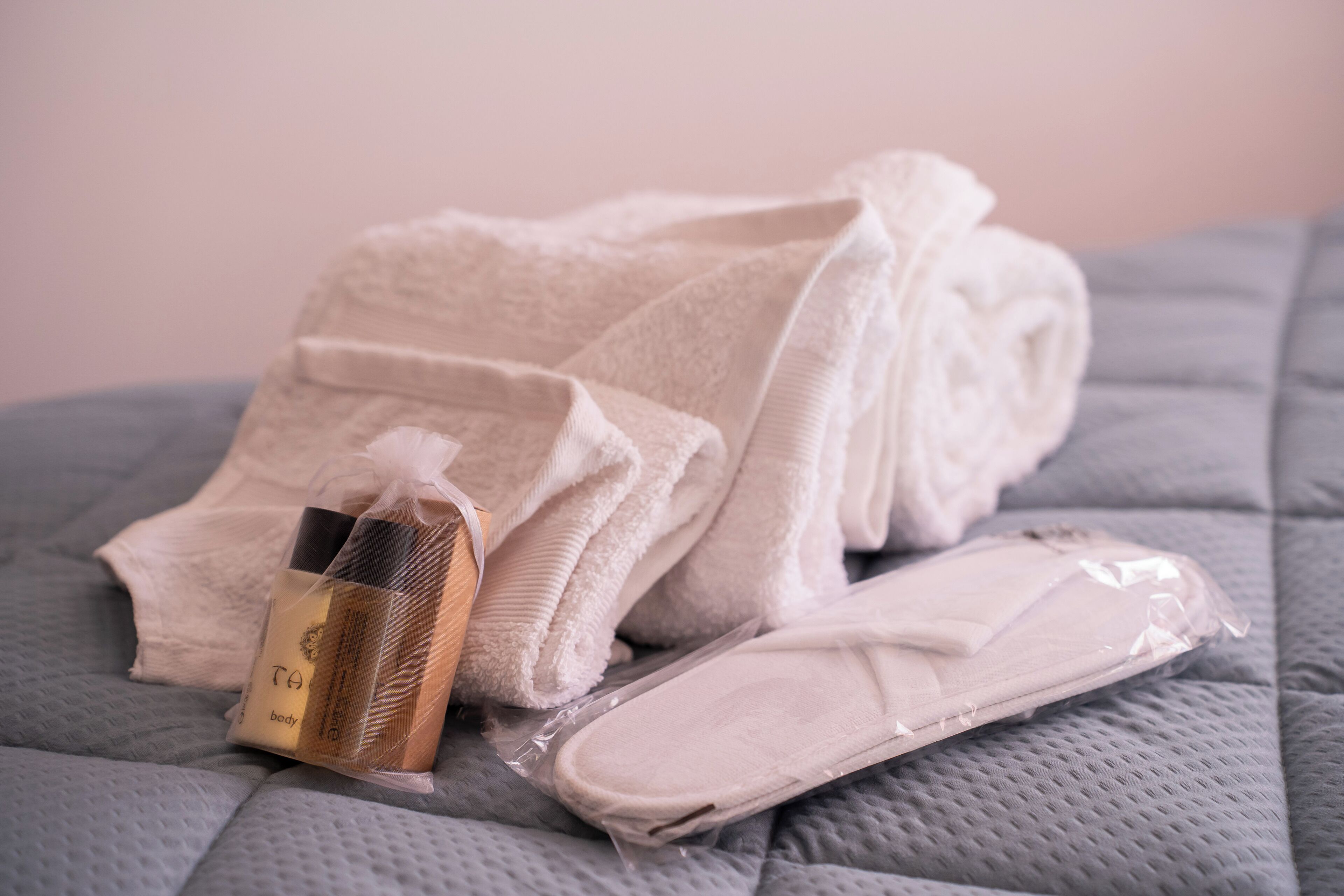 A neatly folded white towel, a bottle of body lotion, and a pair of slippers on a textured gray bedspread.