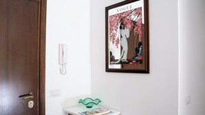 Een kamer met een houten deur, een witte muur, een kunstwerk in een lijst, een wit kastje en een groen voorwerp bovenop.