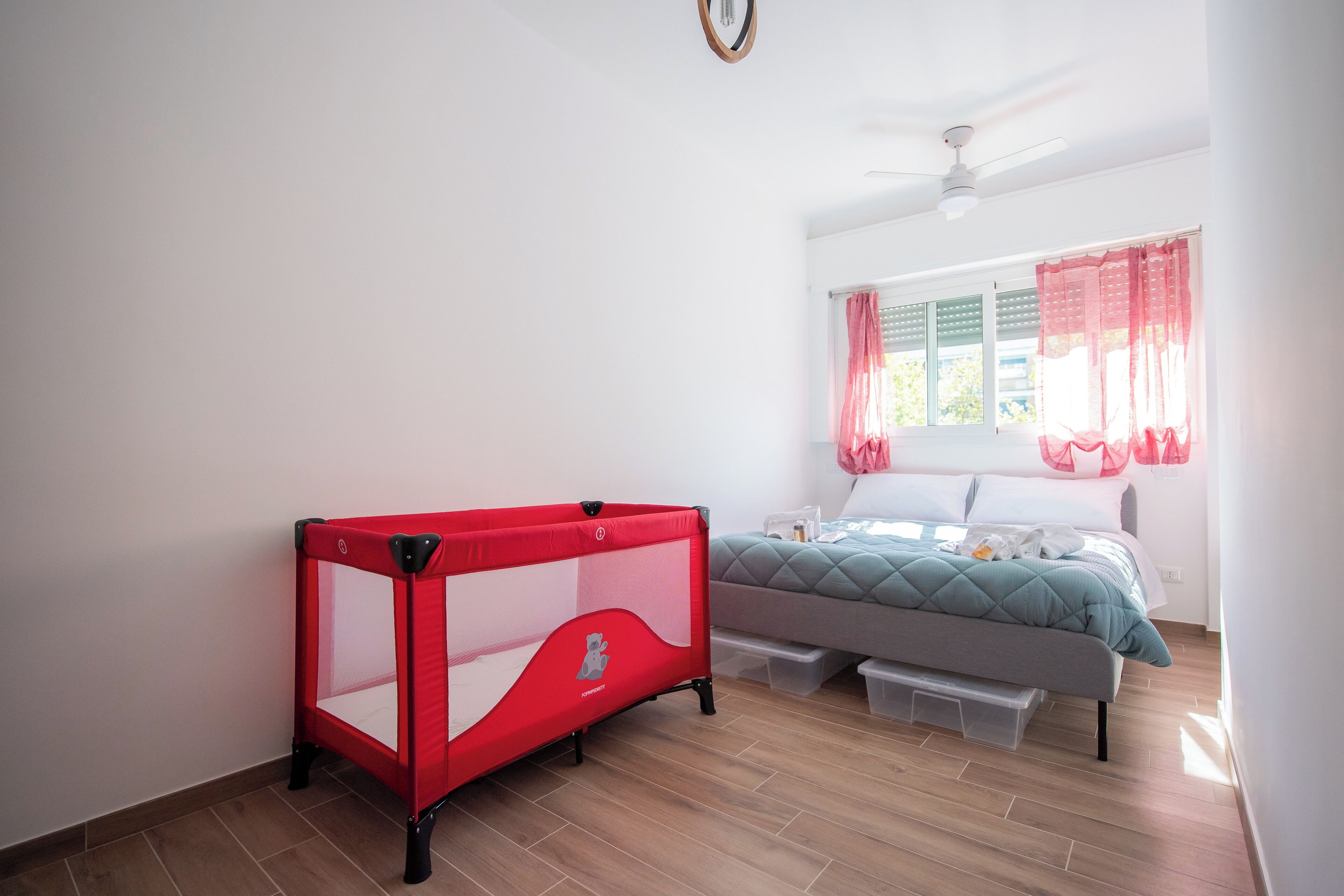 A bedroom with a bed, a red playpen, and a window with curtains.
