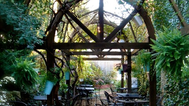 An outdoor café with tables and chairs arranged under a wooden pergola structure, surrounded by lush greenery.