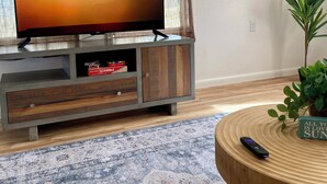 A living room with a flat-screen TV on a wooden cabinet, a patterned rug, a round wooden coffee table, and a potted plant.