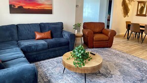 A living room with a blue sofa, a brown leather armchair, a wooden coffee table with a plant, and a large painting of a sunset.