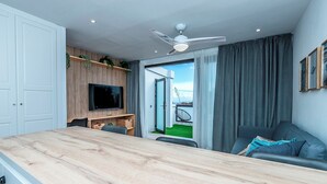 Ein modernes Wohnzimmer mit einem Holzesstisch, einem an einer Holzwand montierten Fernseher, einem Deckenventilator und Blick auf einen Balkon mit einem Fahrrad.