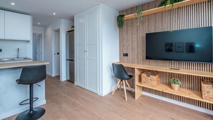 Eine moderne Küche mit weißen Schränken, einer Holz Arbeitsplatte und einem integrierten Fernseher an einer Holzwand. Zwei schwarze Barhocker und ein Holzregal mit Körben und Pflanzen sind ebenfalls vorhanden.