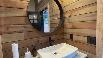 Ein Badezimmer mit Holzwandverkleidung, einem weißen Waschbecken, einem runden Spiegel und zwei Wandleuchten.