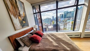 A bedroom with a large window, a bed with a striped blanket, and a wooden nightstand. - Downtown Escape – Walk to Cruise Ships & BC Place | 3 Comfy Beds (Vancouver)