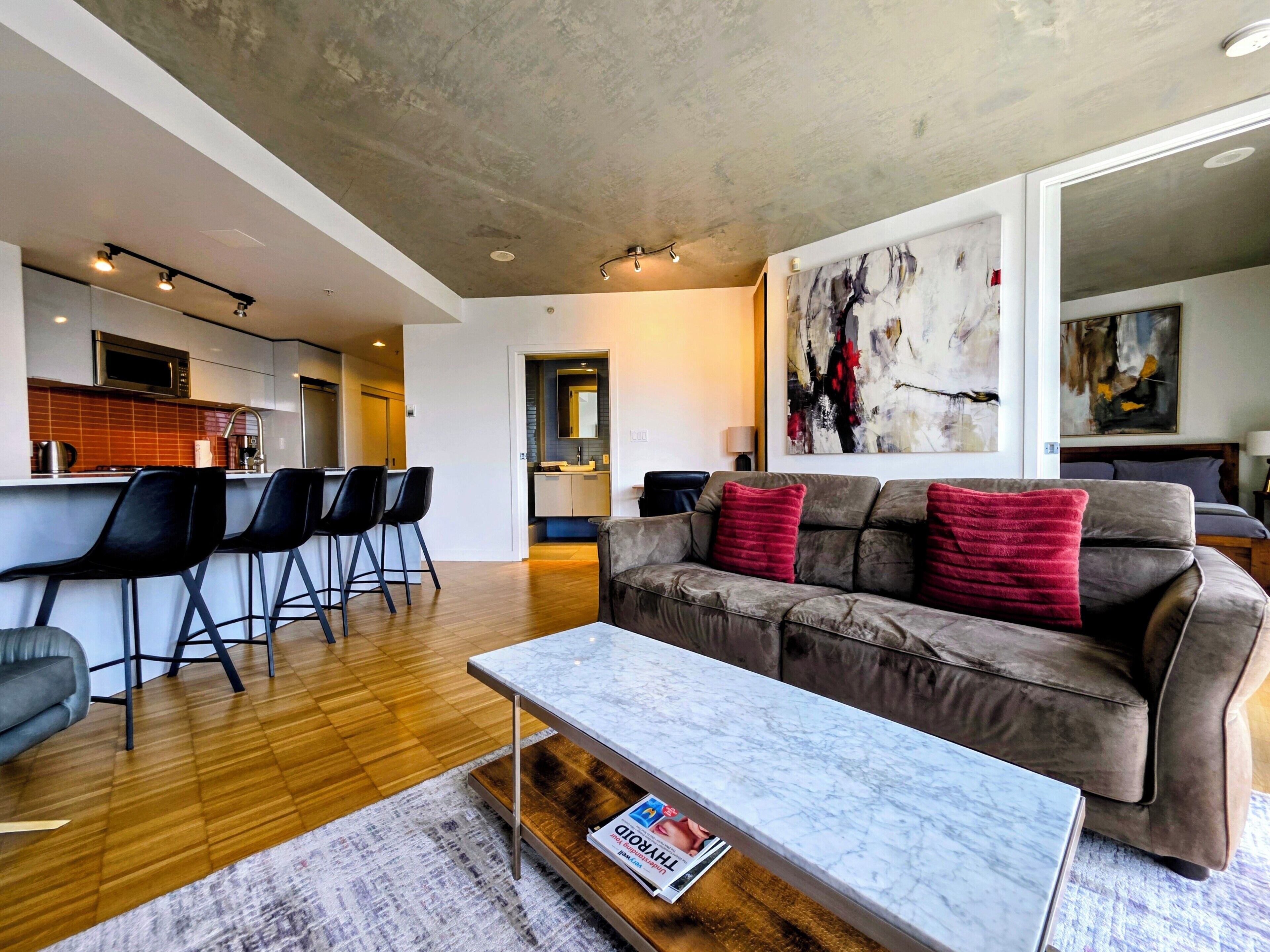 Ein stilvoller Wohnbereich mit einer Couch, einem Couchtisch und Barhocker. Im Hintergrund ist eine Küche erkennbar.