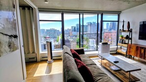 A modern living room with a large window, a sofa, a coffee table, and a bookshelf. - Downtown Escape – Walk to Cruise Ships & BC Place | 3 Comfy Beds (Vancouver)