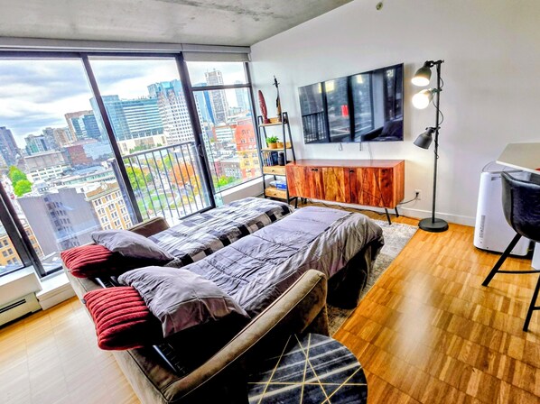 A modern bedroom with a large bed, a TV, a desk, and a view of a cityscape. - Downtown Escape – Walk to Cruise Ships & BC Place | 3 Comfy Beds (Vancouver)
