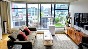 A modern living room with a large window, a sofa, a coffee table, a TV, and a wooden cabinet. - Downtown Escape – Walk to Cruise Ships & BC Place | 3 Comfy Beds (Vancouver)