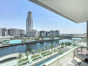 A high-rise hotel with a glass balcony overlooking a river, palm-lined promenade, and a view of a tall building across the water. - Palace Residence - Chic Two Bed with Creek View (Creek Harbour)