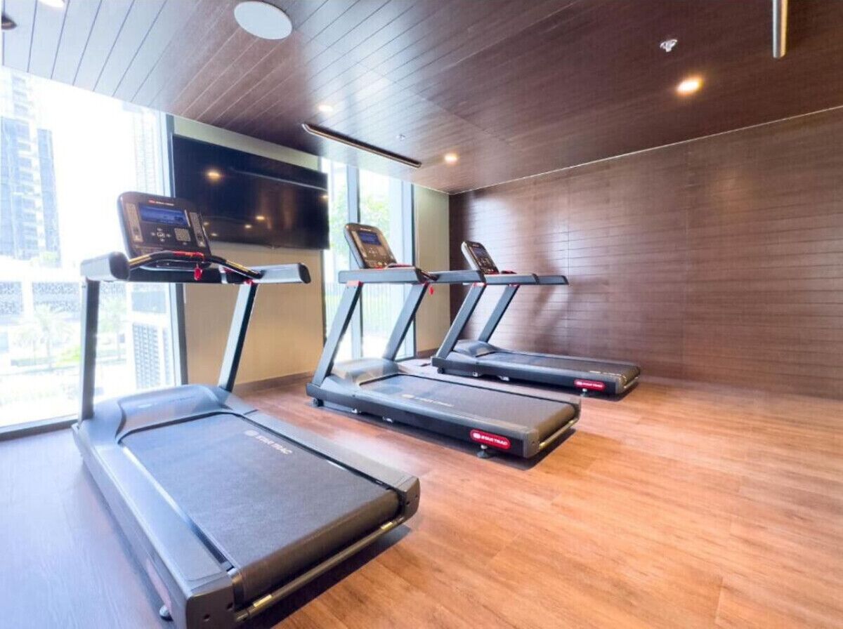 A gym with treadmills and a stationary bike, wooden flooring, and a large window with a city view.