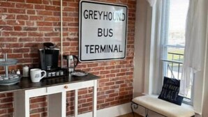 Ein Zimmer mit einer Ziegelwand, einem Schild mit der Aufschrift 'GREYHOUND BUS TERMINAL', einem weißen Schrank, einer Bank an einem Fenster und einer Kaffeemaschine.