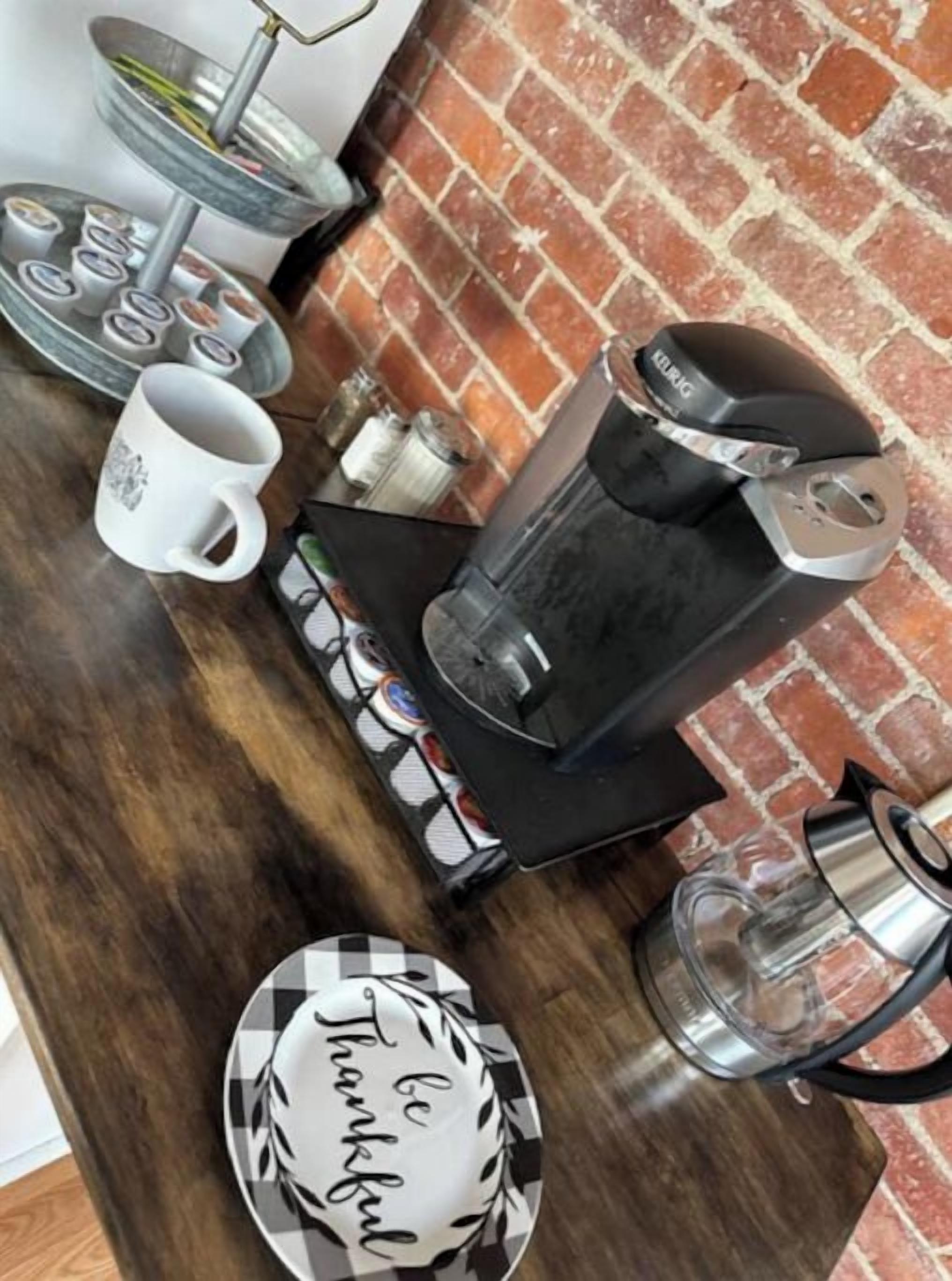 Un taulell de cuina amb una cafetera, una tassa, un plat amb la frase 'be thankful' escrita i una safata amb tasses de cafè.