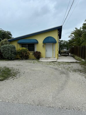 A single-story yellow building with a white door and blue awnings, a gravel driveway, and a boat in the background. - Deep Water Home located in White Marlin Beach Neighborhood (Islamorada)