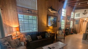 A cozy room with wooden walls, a sofa, a coffee table, and a chair. There are windows with blinds, a lamp, and a painting on the wall. - Deep Water Home located in White Marlin Beach Neighborhood (Islamorada)