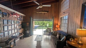 A cozy living room with wooden walls, a ceiling fan, a TV, and a large window overlooking greenery. - Deep Water Home located in White Marlin Beach Neighborhood (Islamorada)