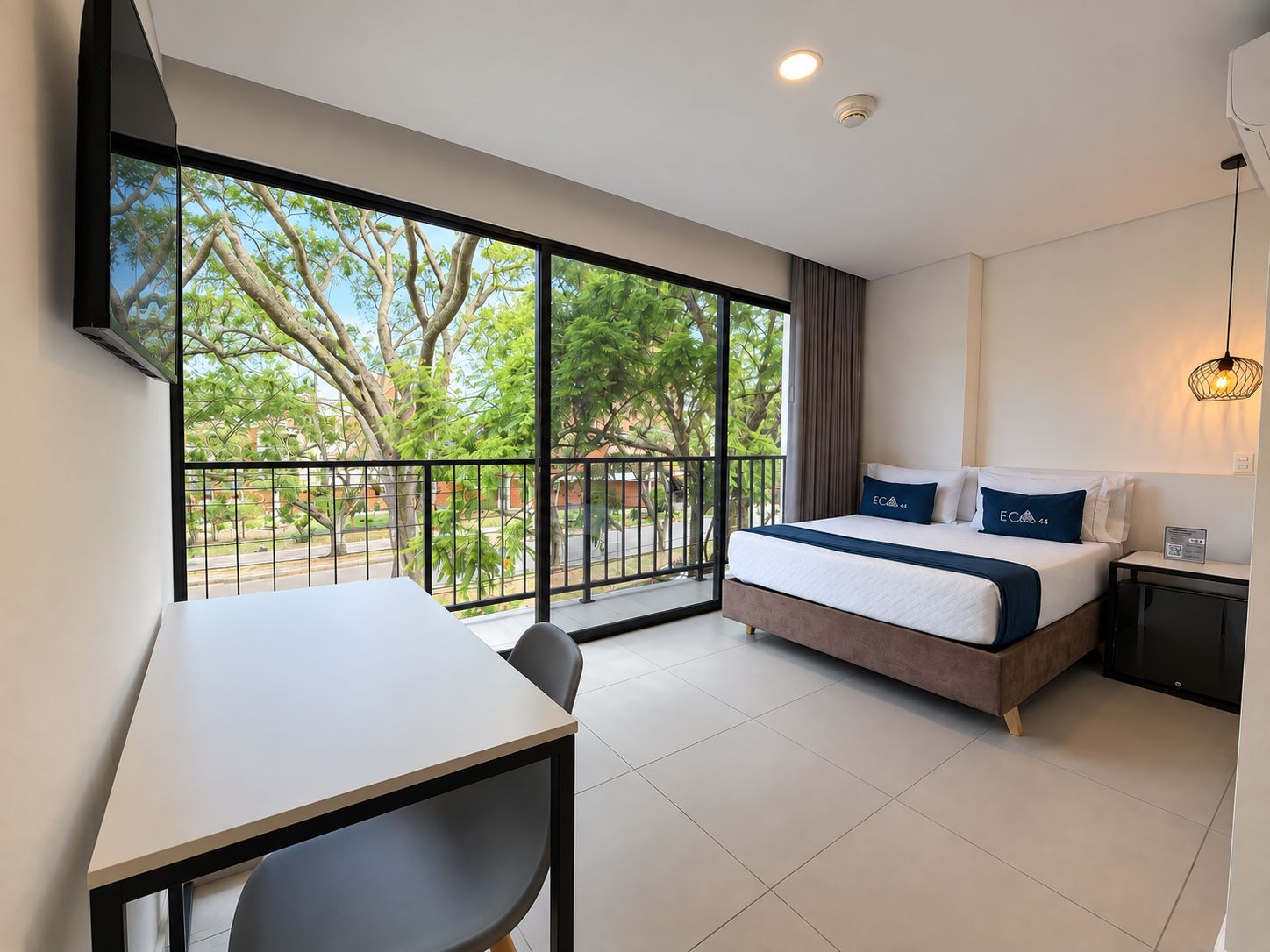 Una habitación de hotel con cama, escritorio y un ventanal con vistas a árboles y una calle.