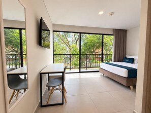 A hotel room with a bed, a desk, a chair, and a large window offering a view of trees and a balcony. - Hotel ECO 44 (Medellín)