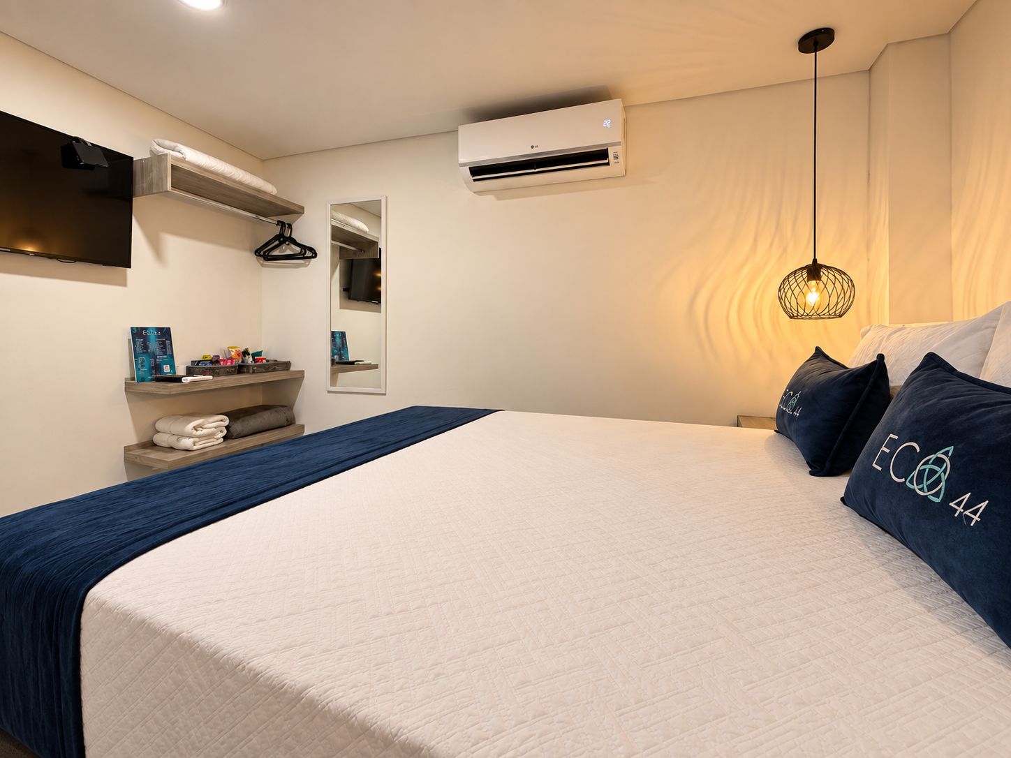 A hotel room with a bed, a hanging light fixture, a wall-mounted air conditioner, and a shelf with towels.