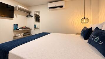 A hotel room with a bed, a hanging light fixture, a wall-mounted air conditioner, and a shelf with towels.