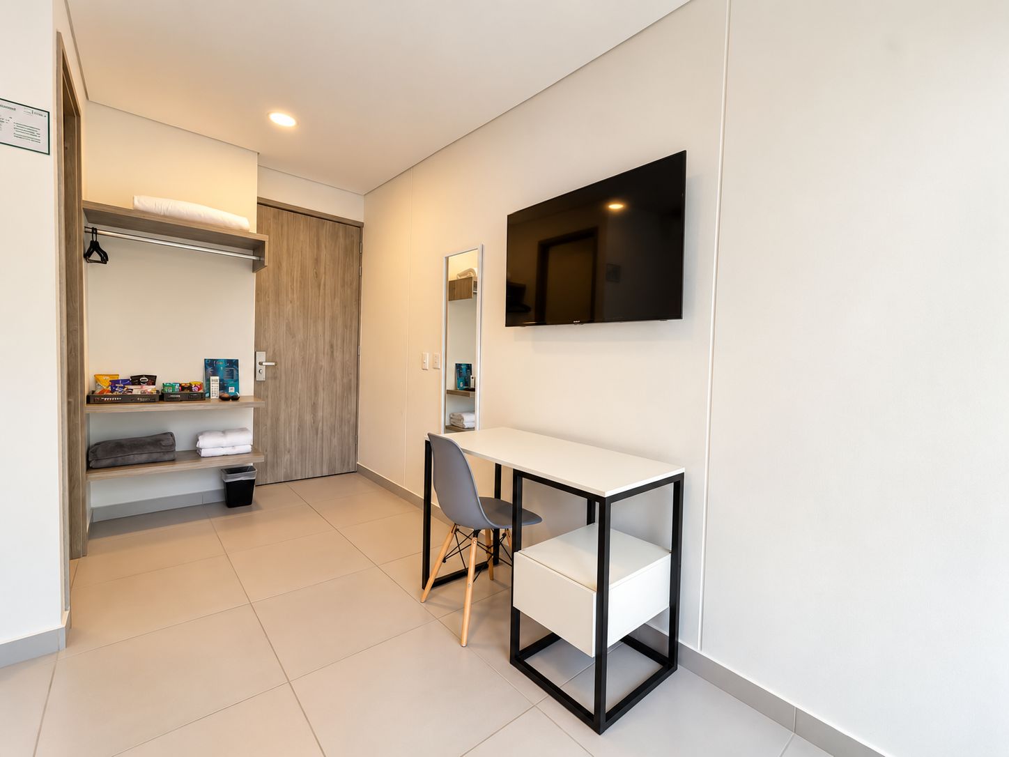 A modern hotel room with a white desk, a chair, a flat-screen TV mounted on the wall, and a wooden wardrobe.