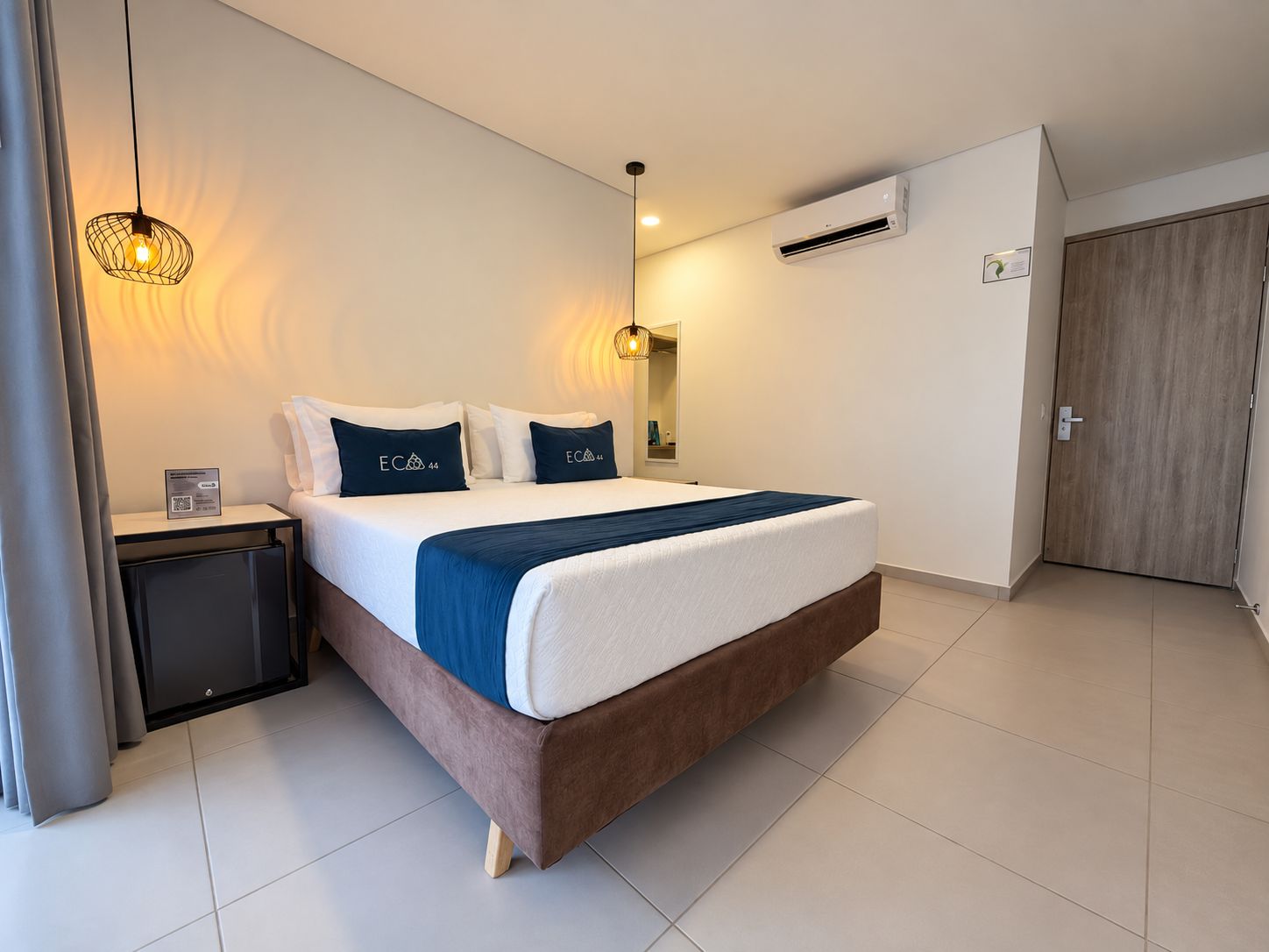 A modern hotel room with a bed, bedside tables, an air conditioning unit, and a door.