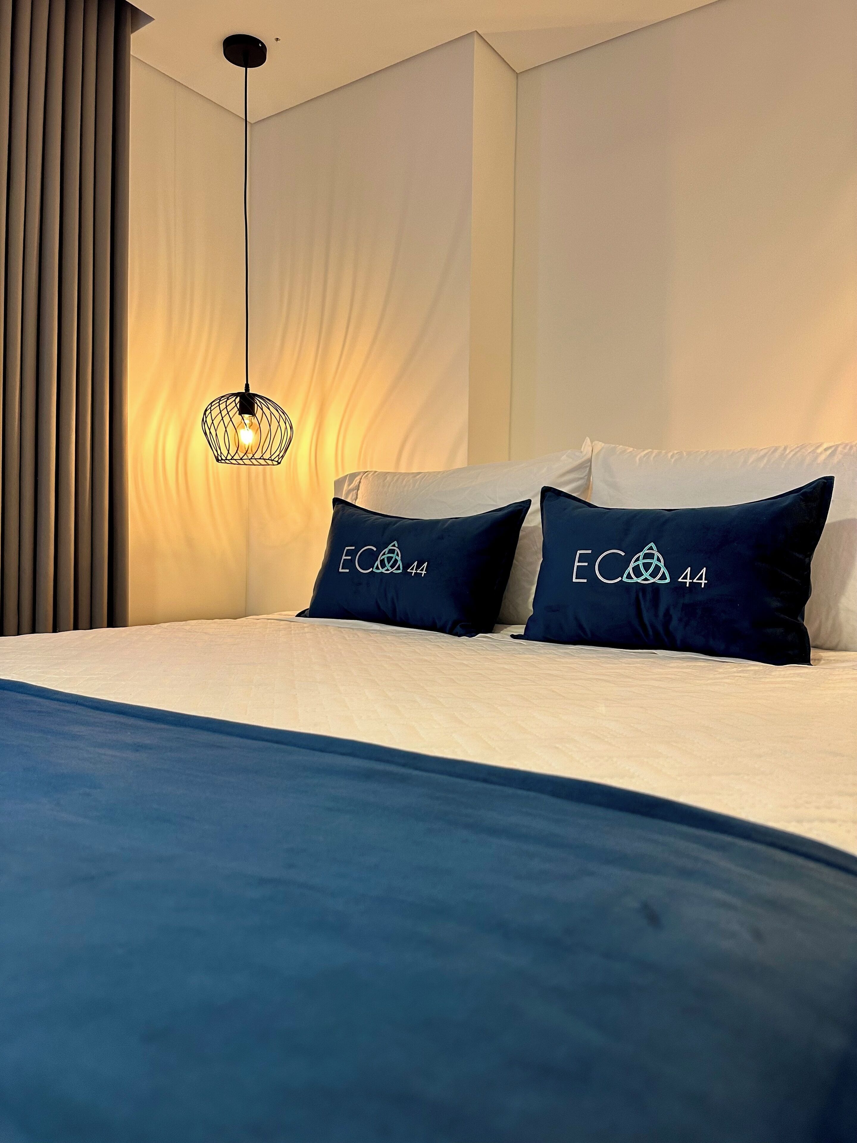 A modern bedroom with a bed, two pillows with a logo, a hanging light fixture, and a curtain.