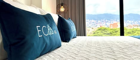 A bed with white bedding and two teal pillows with embroidered text. A cityscape is visible through a window.