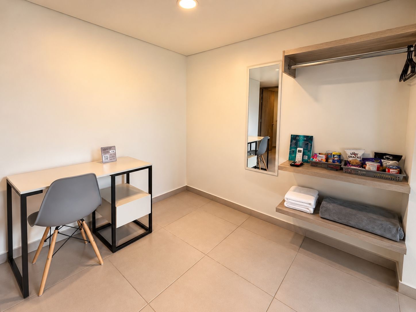 Ein modernes Zimmer mit Schreibtisch, Stuhl, Spiegel und Regalen mit Snacks und Pflegeprodukten.