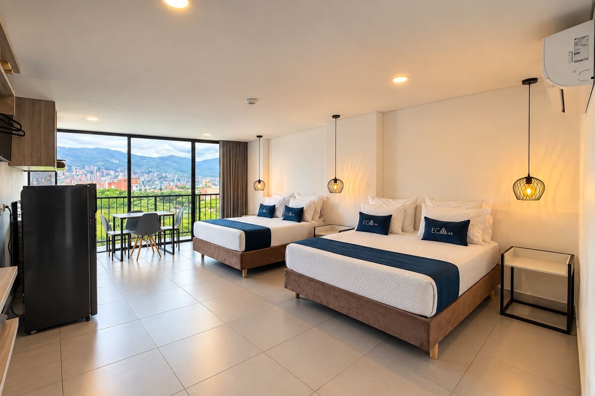 A hotel room with two beds, a dining area, and a view of a cityscape.