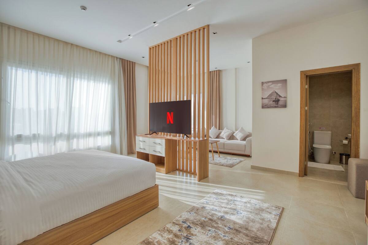 A modern hotel room with a large bed, a TV, a wooden partition, a sofa, and a bathroom visible through an open door.
