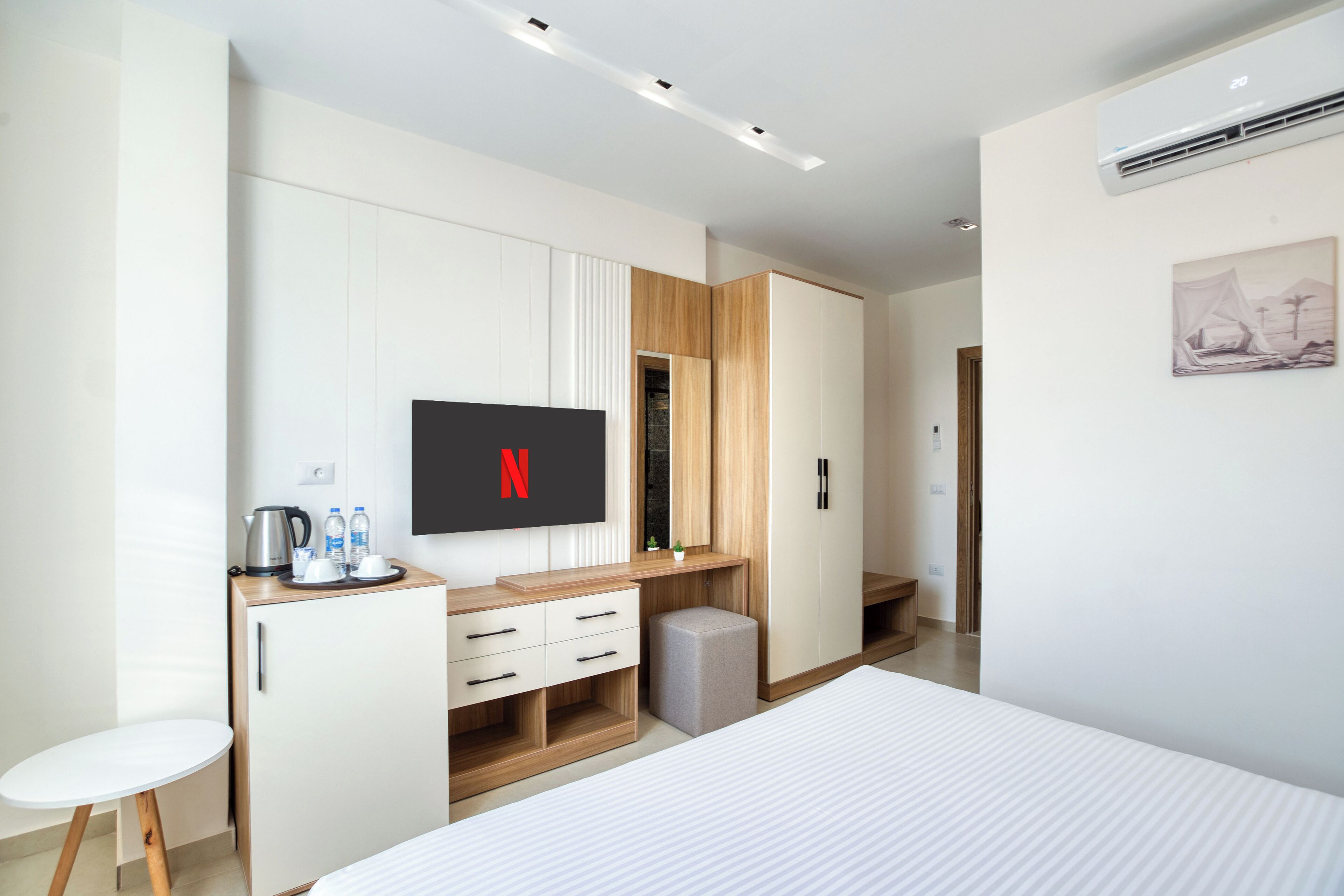 A modern hotel room with a bed, a TV mounted on the wall, a desk with a kettle, and a small table.