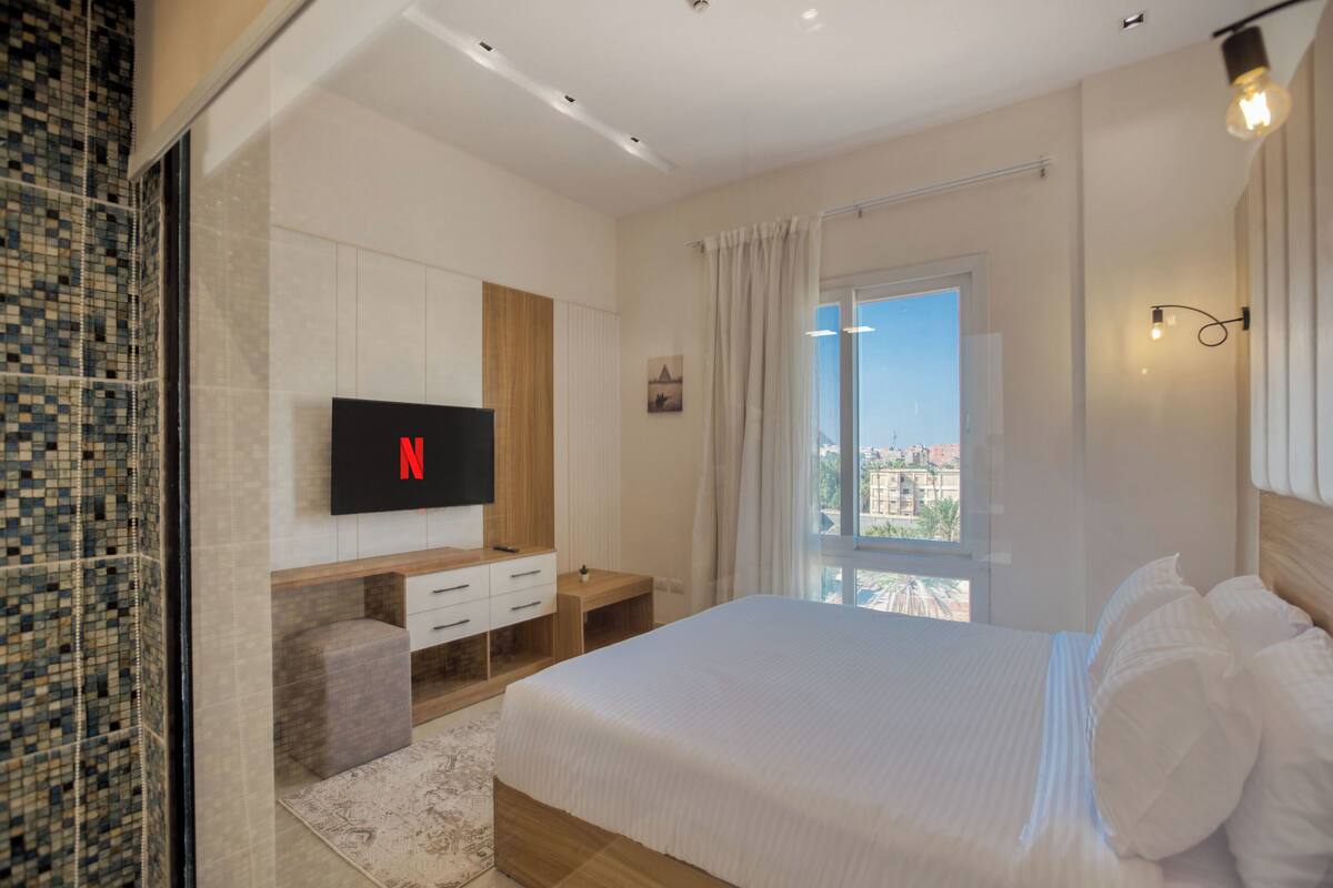 A modern hotel room with a large bed, a TV mounted on the wall, a small desk, and a window with a city view.