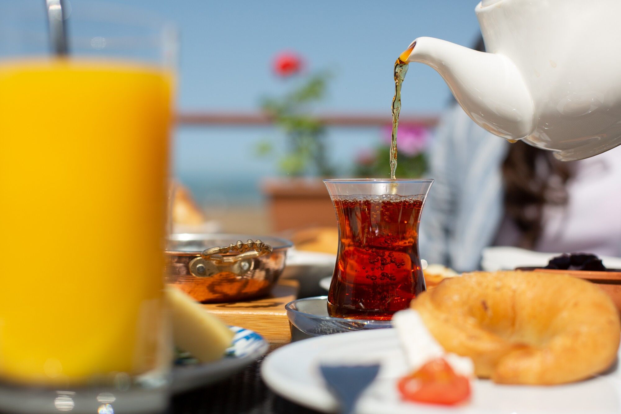 A breakfast setting with a teapot pouring tea into a glass, a croissant, and a plate with a tomato.