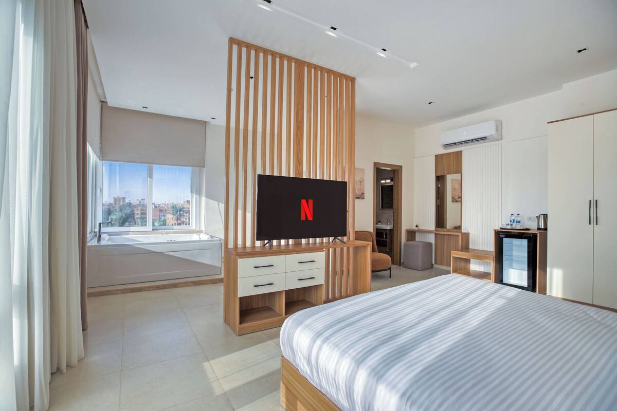 A modern hotel room with a large bed, a TV mounted on a wooden panel, a desk, and a wardrobe.
