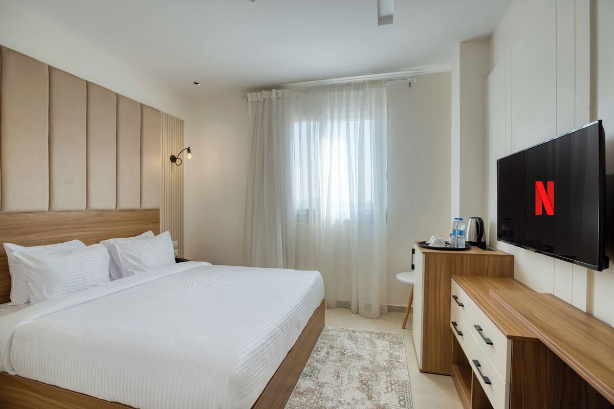 A hotel room with a large bed, a wooden headboard, a flat-screen TV, a bedside table with a kettle, and a window with sheer curtains.