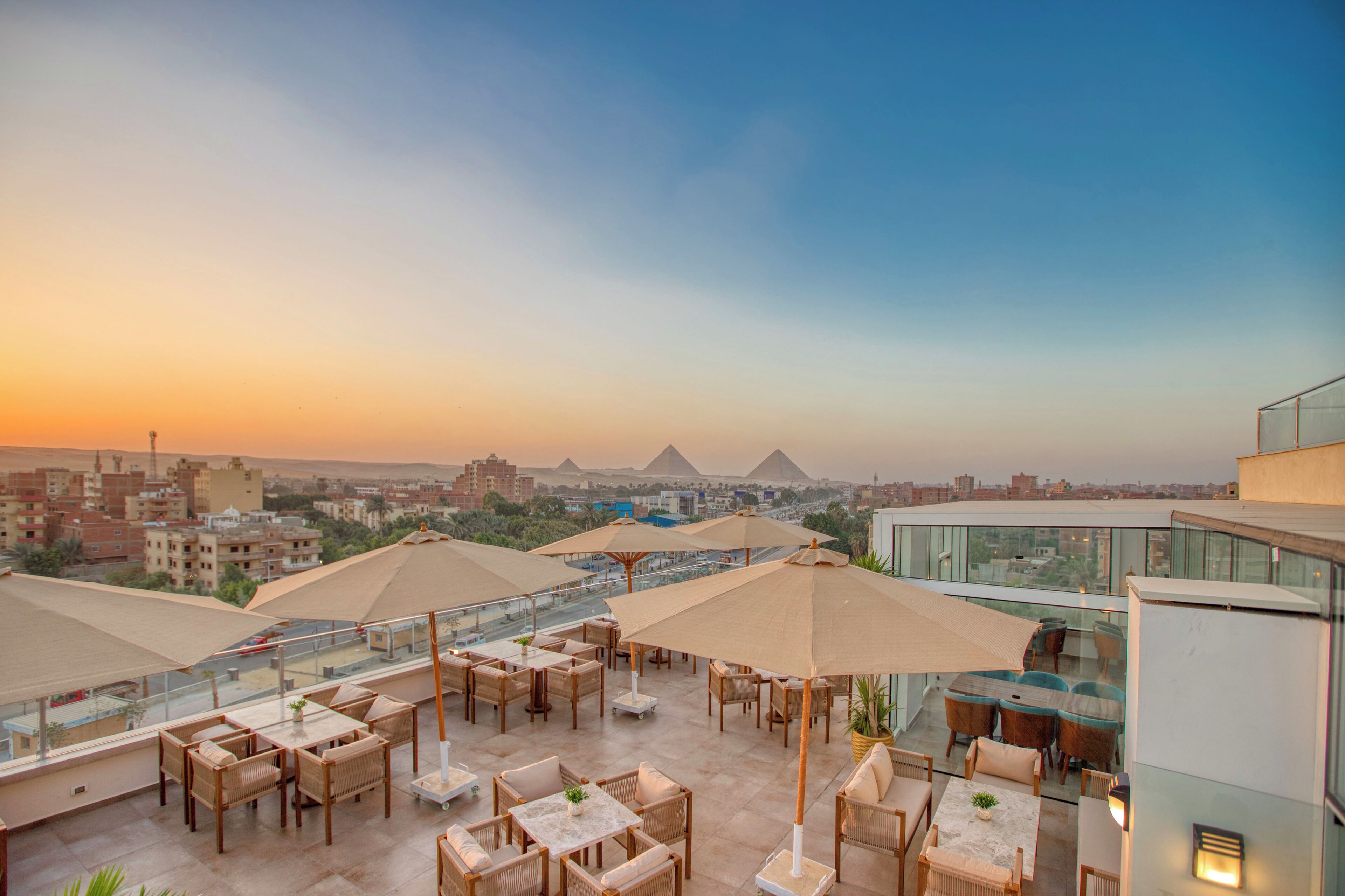 A rooftop terrace with outdoor seating and umbrellas, overlooking a cityscape and pyramids.