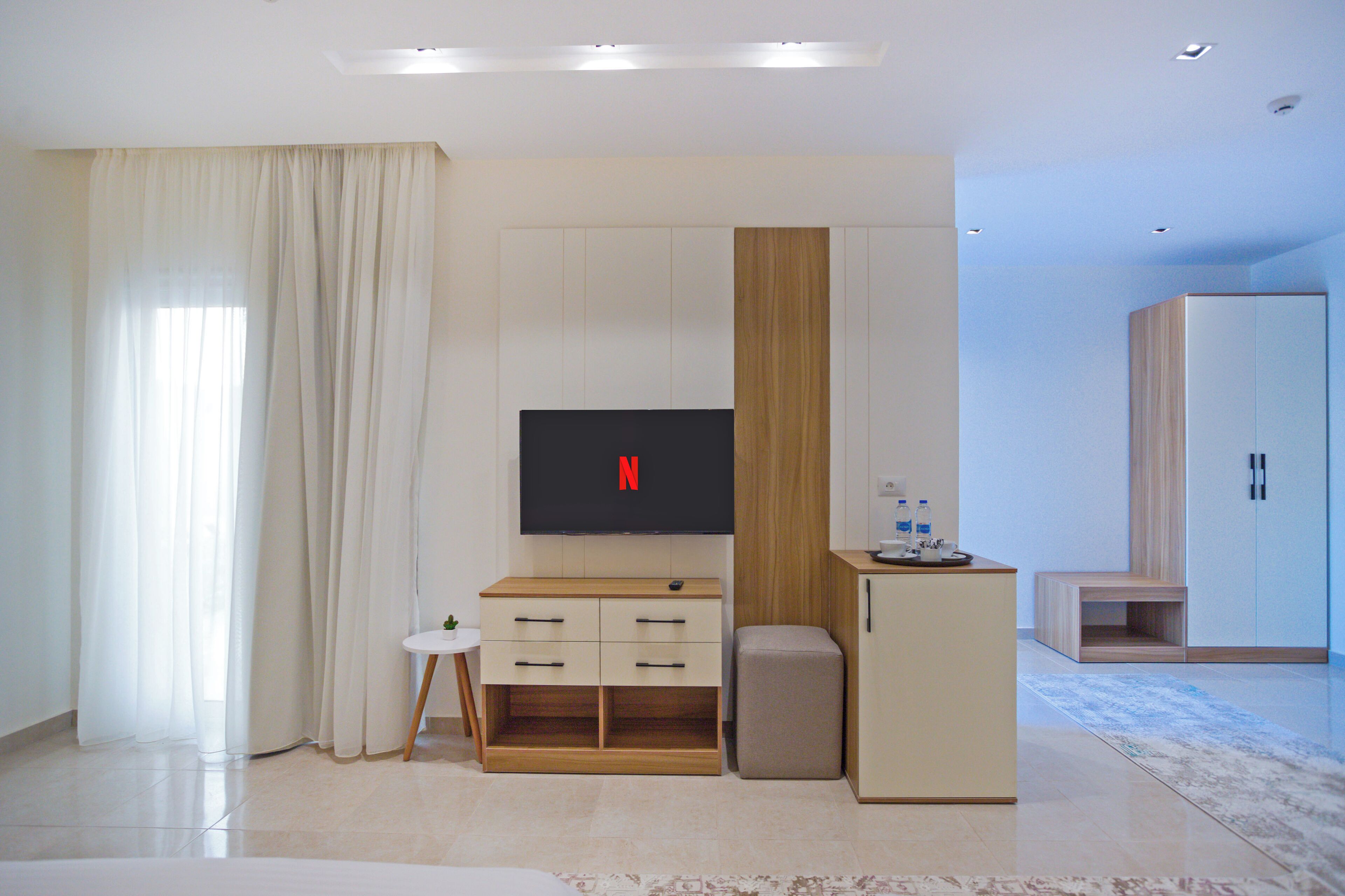 A modern hotel room with a flat-screen TV mounted on the wall, a small wooden side table with a remote control, a beige sofa, and a wardrobe.