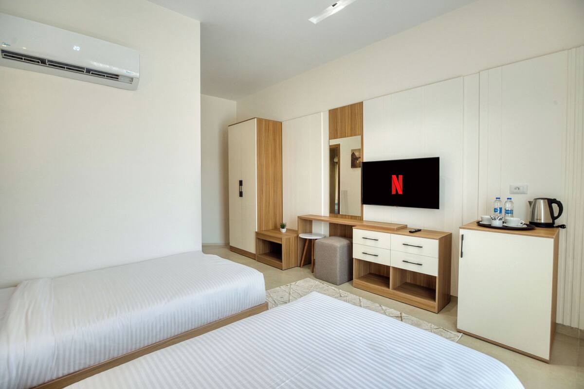 A modern hotel room with two single beds, a wooden wardrobe, a desk with a mirror, a television, and a small kitchenette area.
