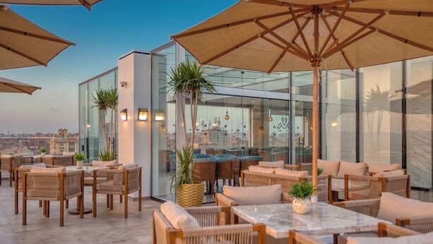 A rooftop terrace with outdoor seating, large umbrellas, and glass walls offering city views. - Mazar Pyramids Hotel (Giza)