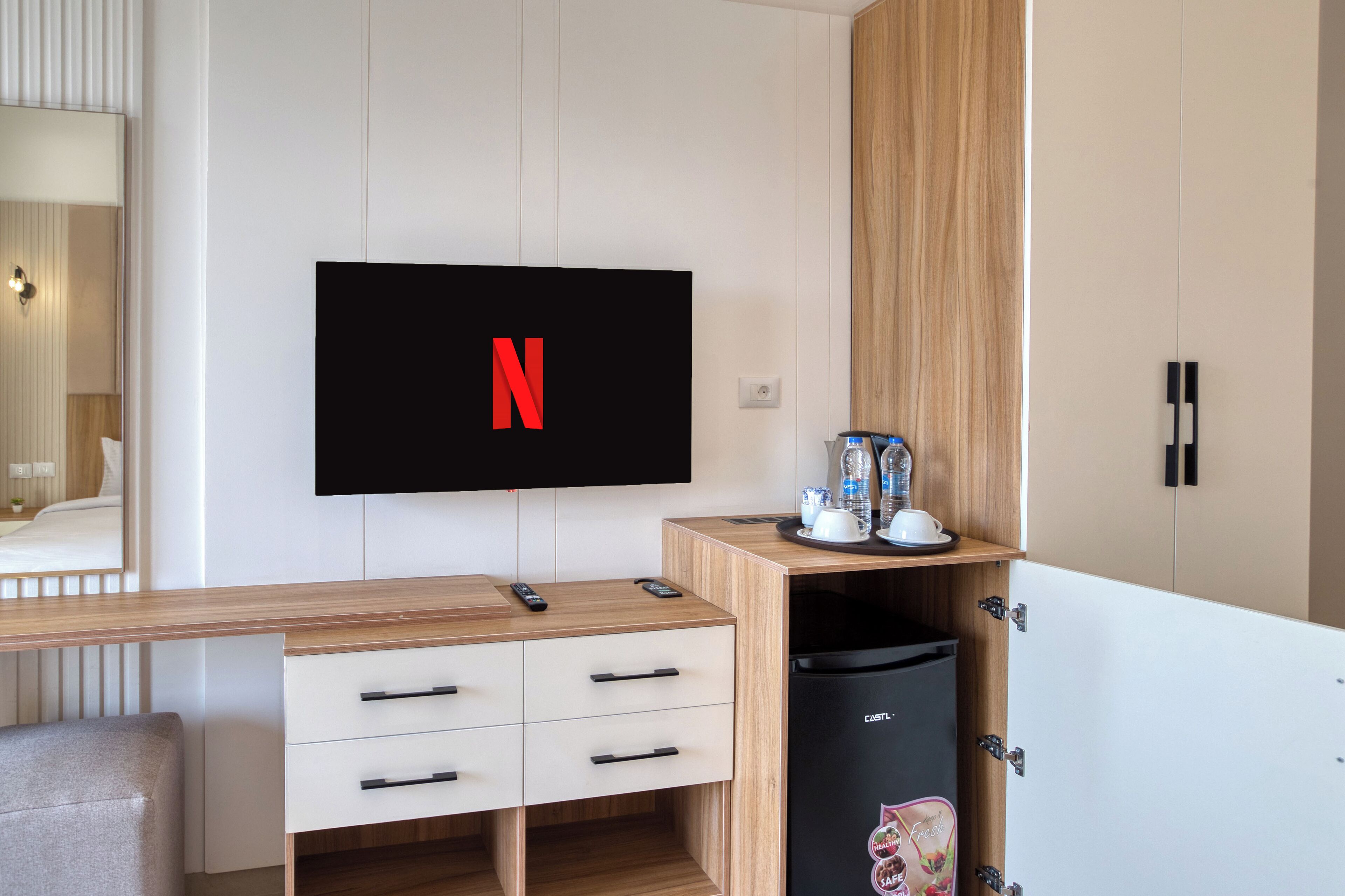 A modern hotel room with a wooden desk, a flat-screen TV mounted on the wall, a minibar, and a wardrobe with a mirror.