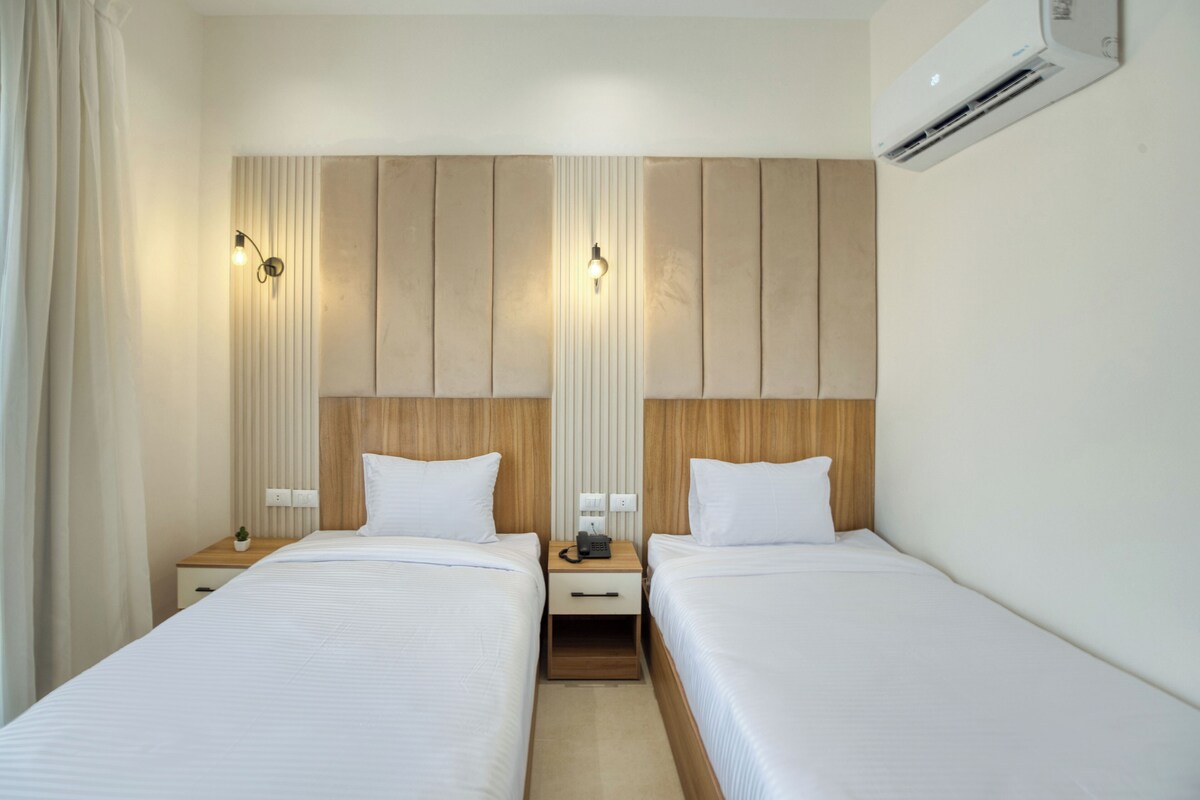 Une chambre d’hôtel avec deux lits simples, une tête de lit en bois, une table de chevet avec un téléphone et un appareil de climatisation.