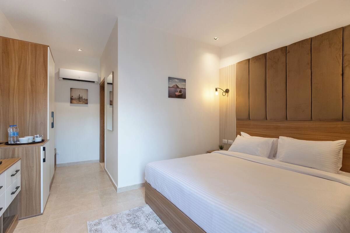 A modern hotel room with a wooden headboard, a large bed, a desk with a chair, and a small kitchenette.