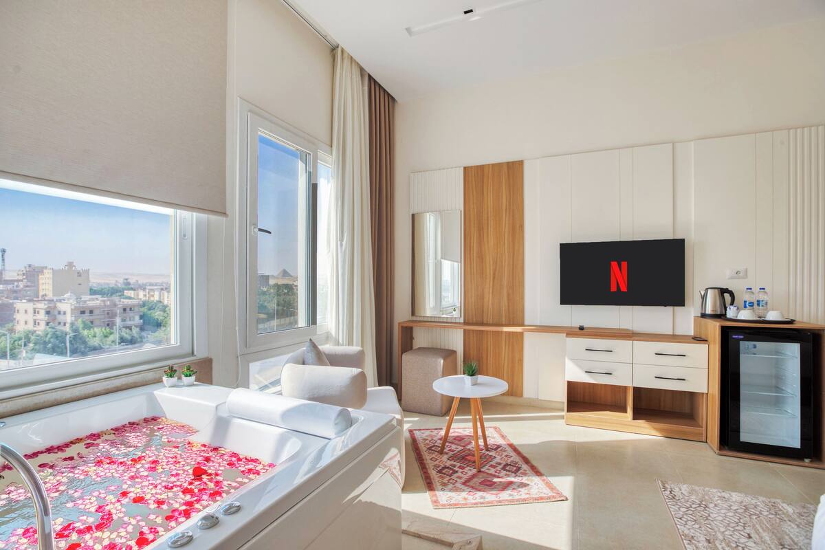A modern hotel room with a large window, a bathtub with rose petals, a wooden desk, a television, and a minibar.