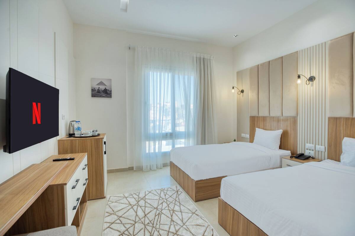 A hotel room with two beds, a TV mounted on the wall, a wooden desk, and a bedside table with a phone and lamp.