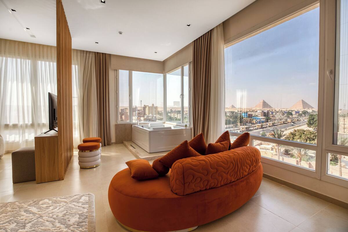 A modern living room with a large window offering a city view, a cozy orange sofa, and a flat-screen TV.